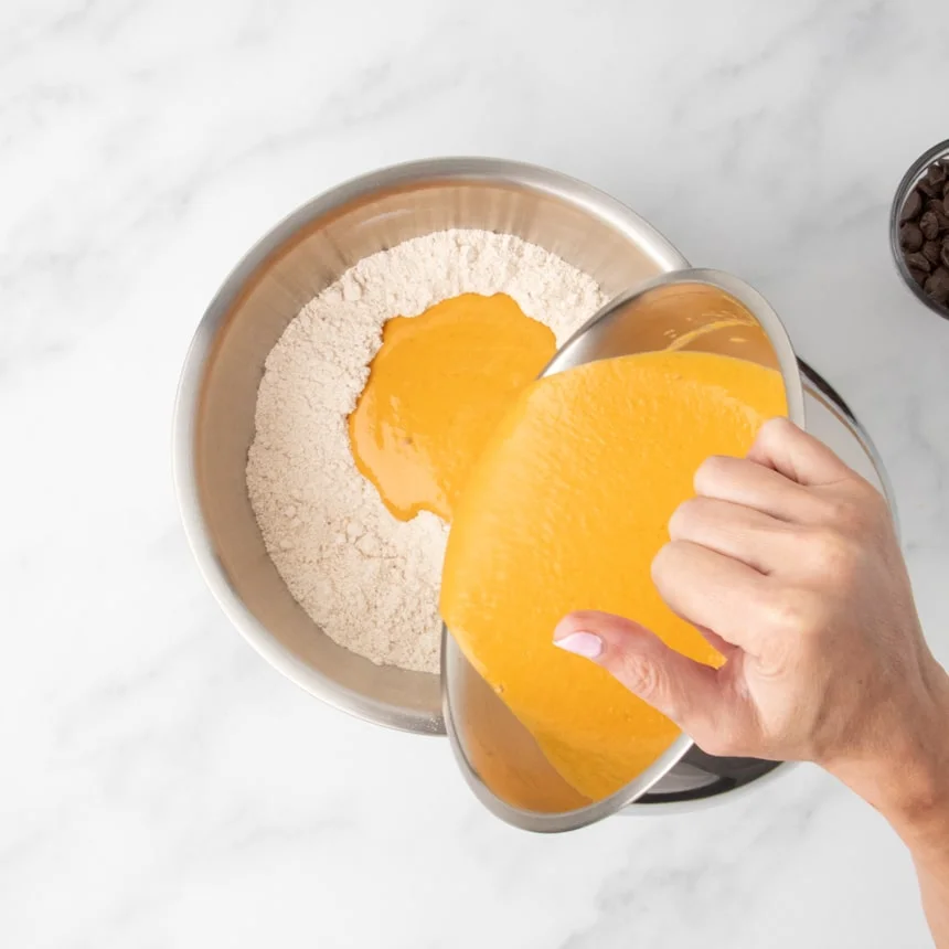 A metal bowl holding an orange mixture is being poured into another metal bowl with a light-colored powder