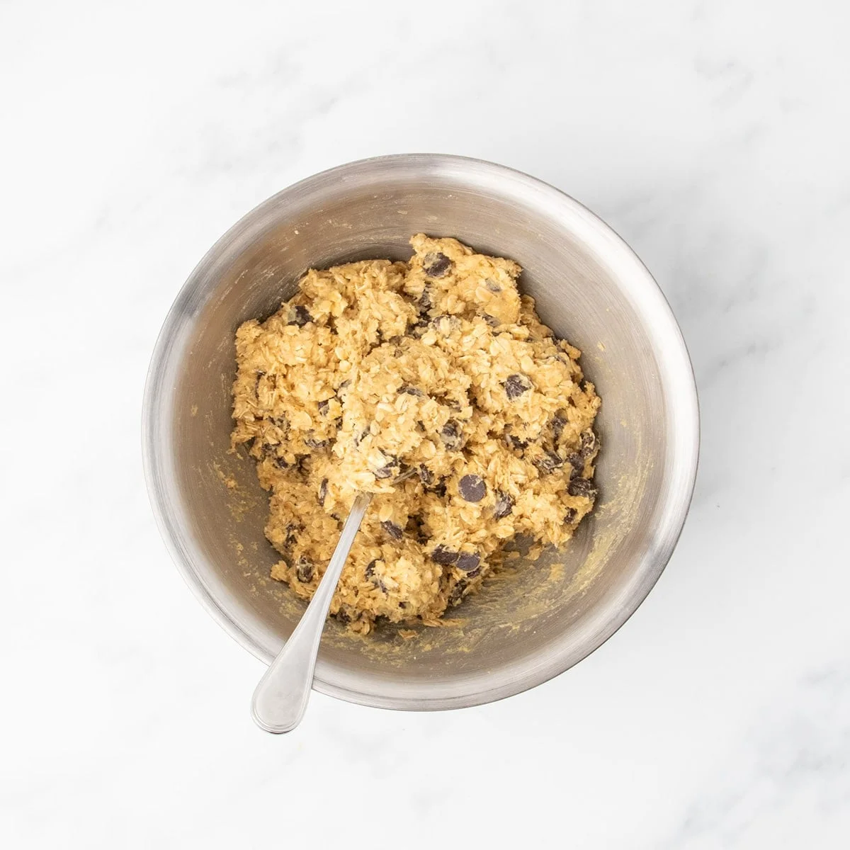 A metal mixing bowl filled with raw thick oatmeal cookie dough with visible chocolate chips, and a metal spoon in the bowl.