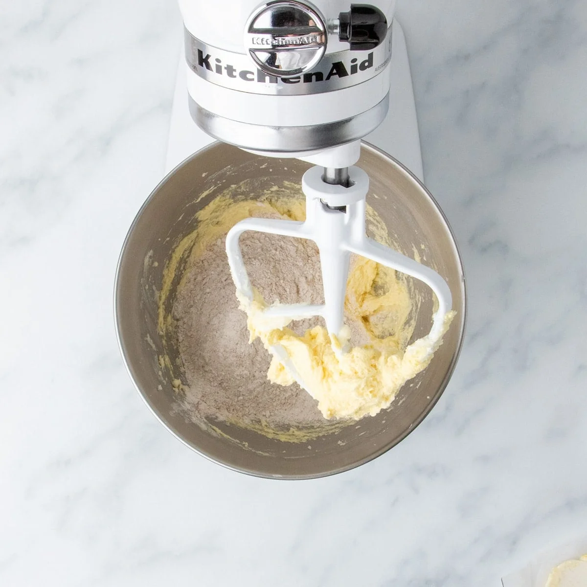 A metal mixing bowl in stand mixer with egg, butter and sugar mixture on lifted paddle. 