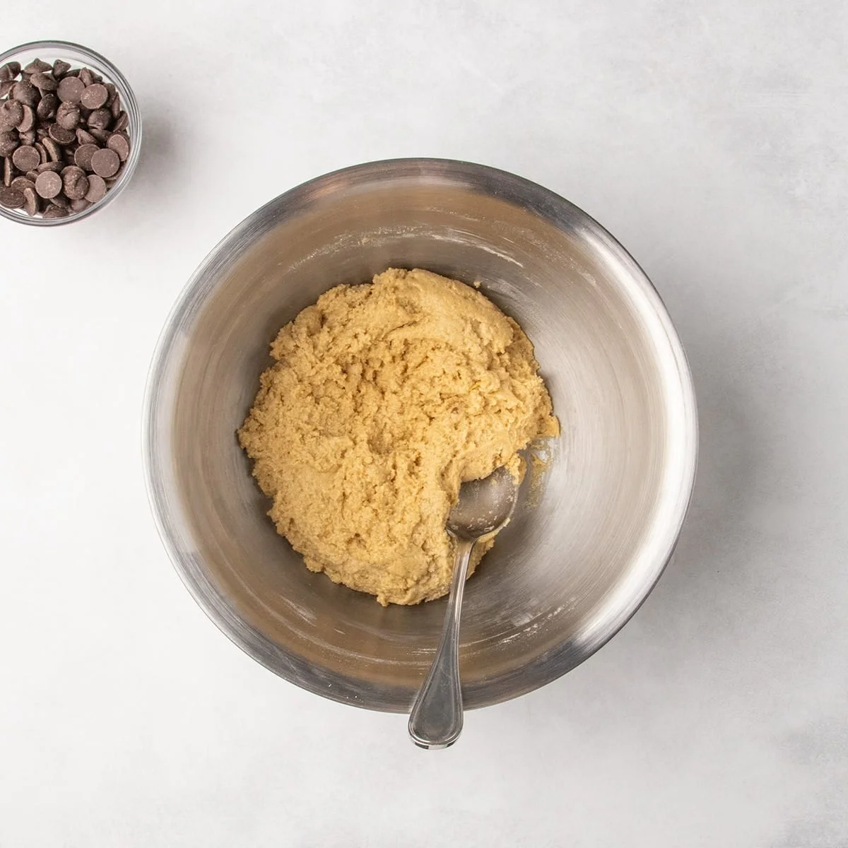 A metal mixing bowl with thick light brown cookie dough and a spoon inside, with a bowl of chocolate chips nearby.