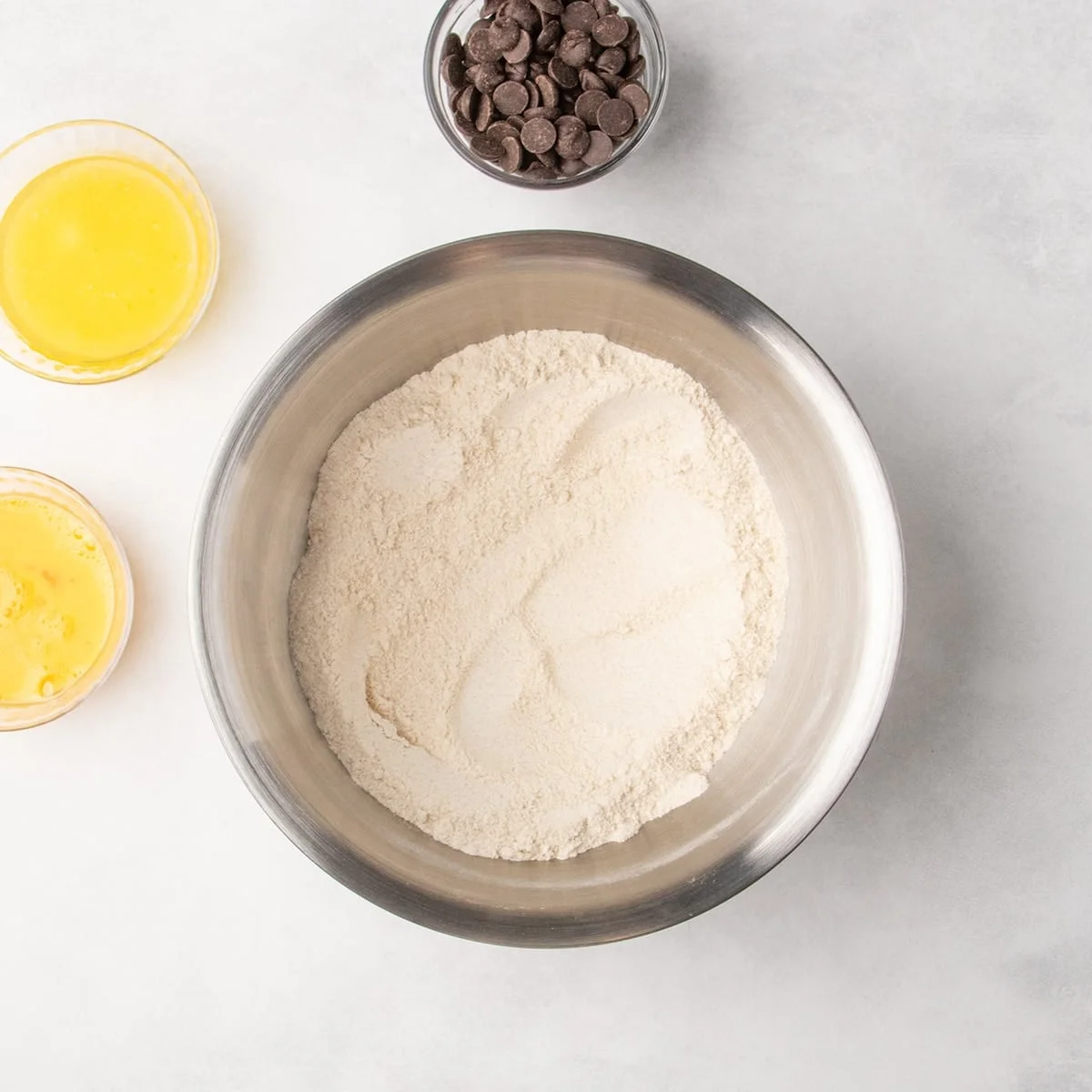 A metal mixing bowl with whisked dry ingredients, with bowls of yellow liquid and chocolate chips nearby.