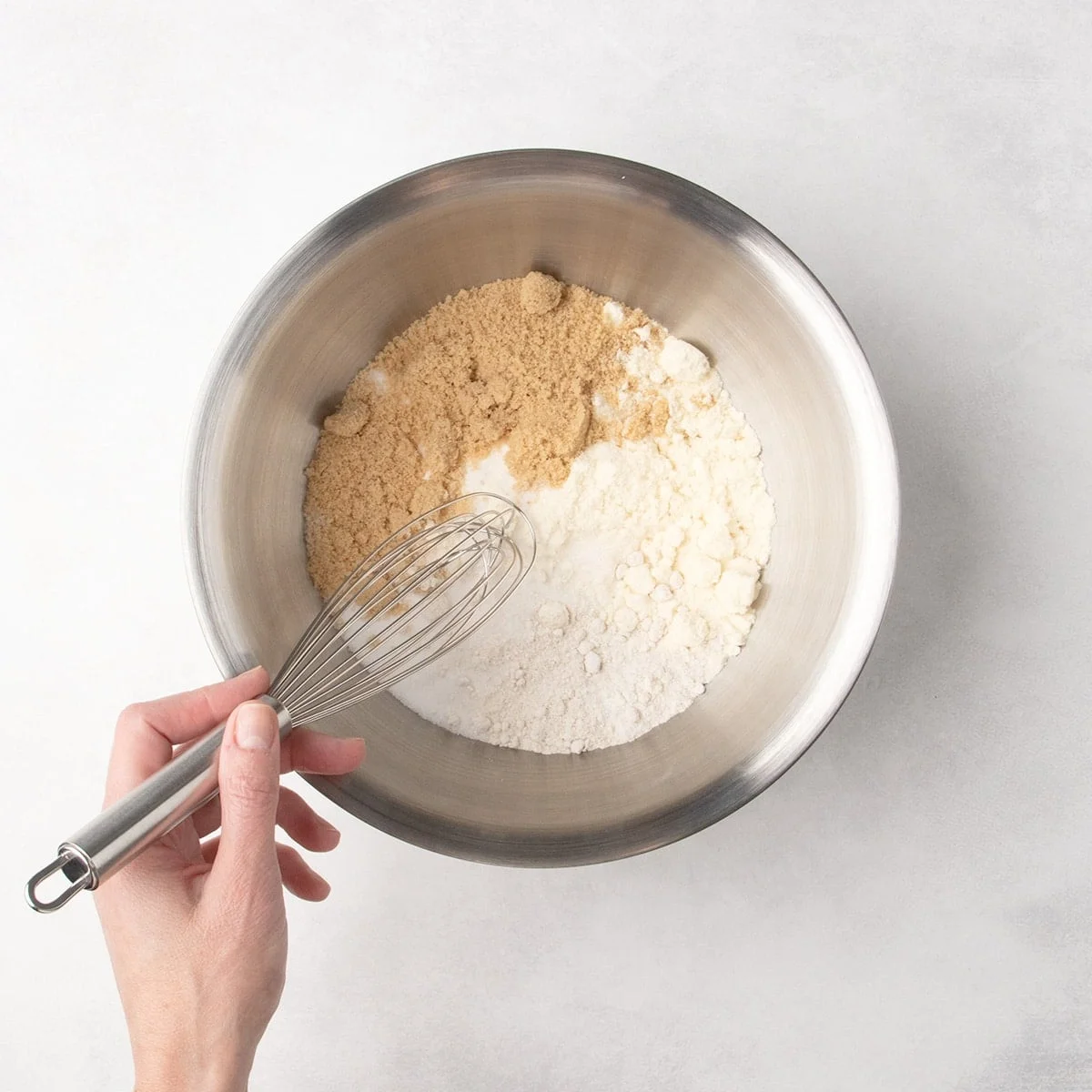 A metal mixing bowl with white and brown dry ingredients and a hand holding a whisk about to mix the contents of the bowl.
