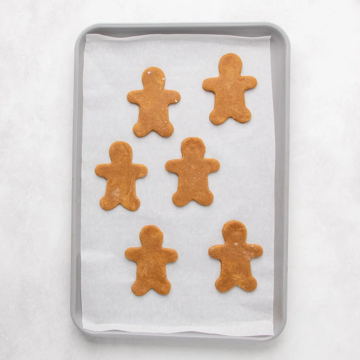 A parchment-lined baking sheet with six unbaked brown gingerbread-person cookies arranged in two columns.