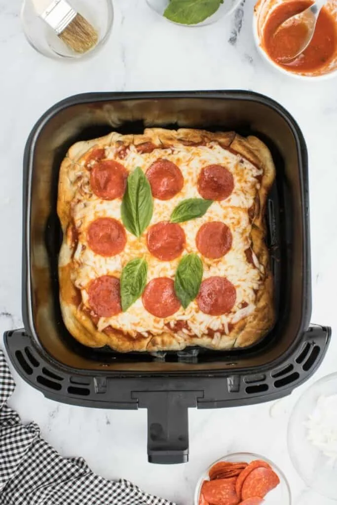 A pepperoni pizza baked in the air fryer, topped with basil, resting in a black air fryer basket.