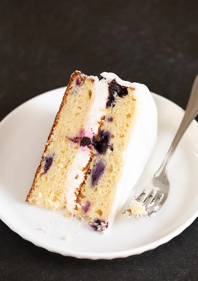 A piece of gluten free lemon blueberry cake with white frosting, on a plate.