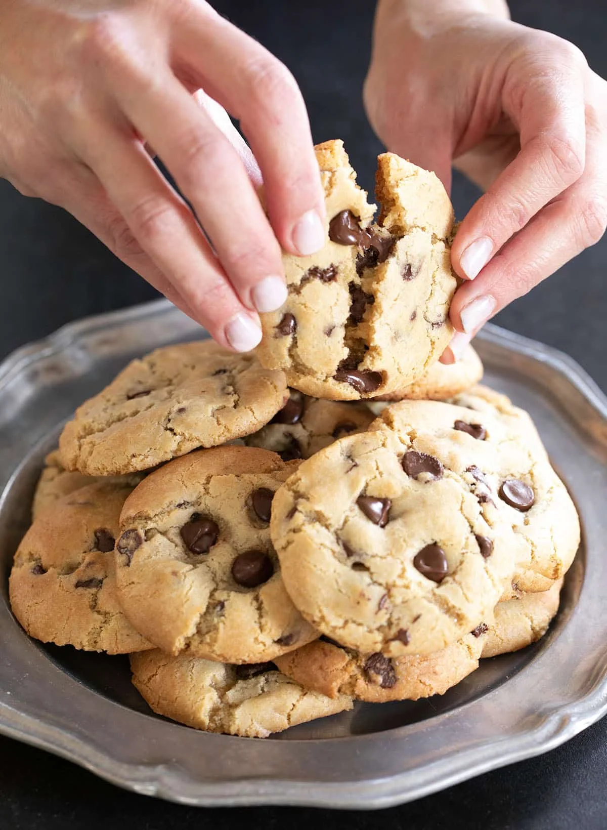 A pile of the cookies on a plate with a hands breaking one open.