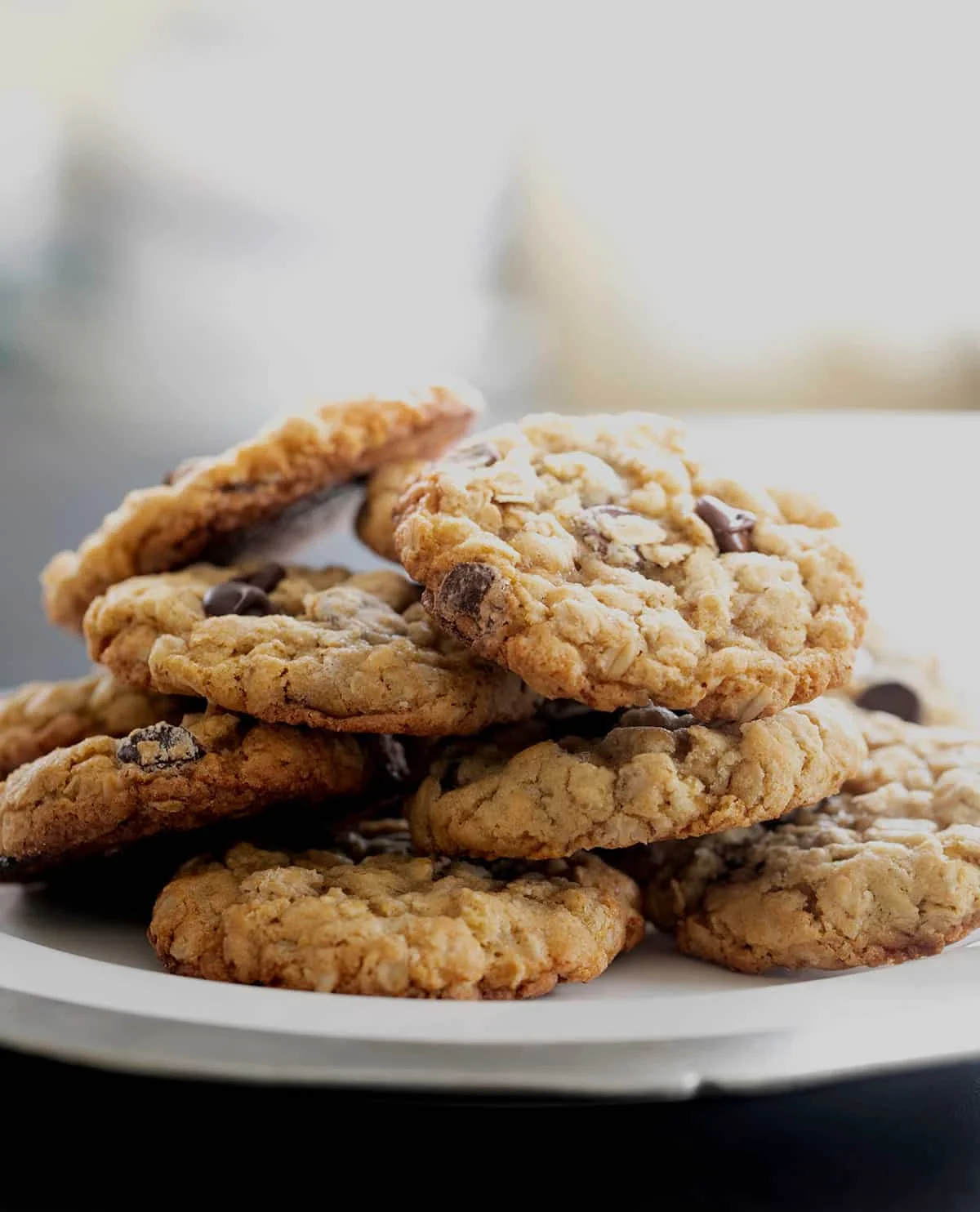 A plate of 8 baked cookies with oats and chocolate chips in a haphazard pile on a white serving platter with a metal edge.