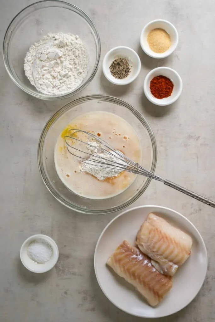 A plate of cod, several small bowls with individual ingredients to prepare fish batter, one larger bowl with the fish batter ingredients.