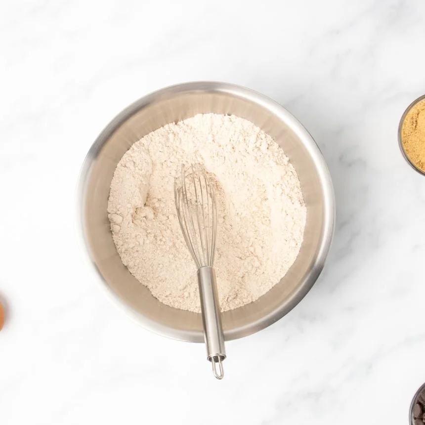 A round metal mixing bowl holds a dry, light-colored powder and a whisk