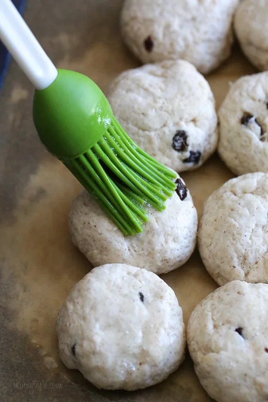 A rubber basting brush brushing egg wash over balls of hot cross bun dough.