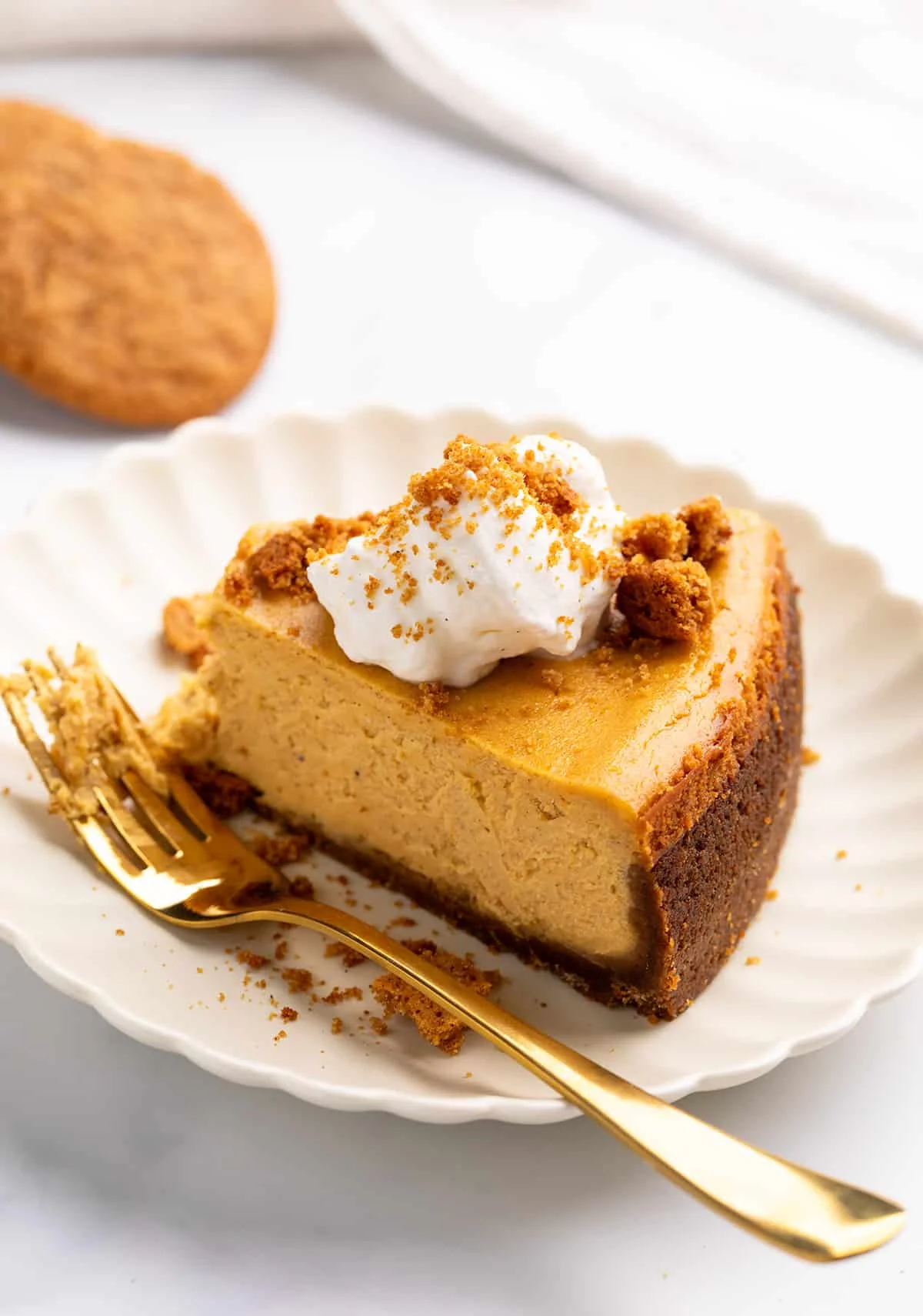 A side view of a slice of pumpkin spice cheesecake with a dark, thick crust, garnished with a dollop of whipped cream and cookie crumbles.