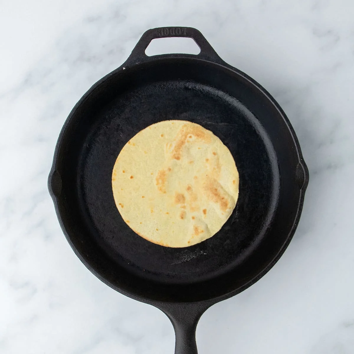 A single tortilla cooking in a black cast iron skillet on a marble surface.