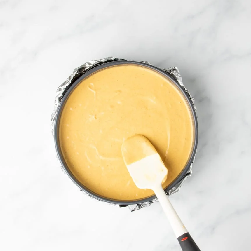 A spatula spreading the smooth pumpkin cheesecake filling evenly over the cookie crust in a springform pan before baking.