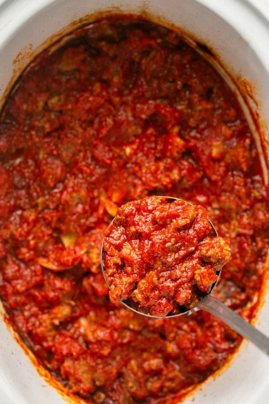 A spoon lifting sausage pasta sauce from a crock pot.