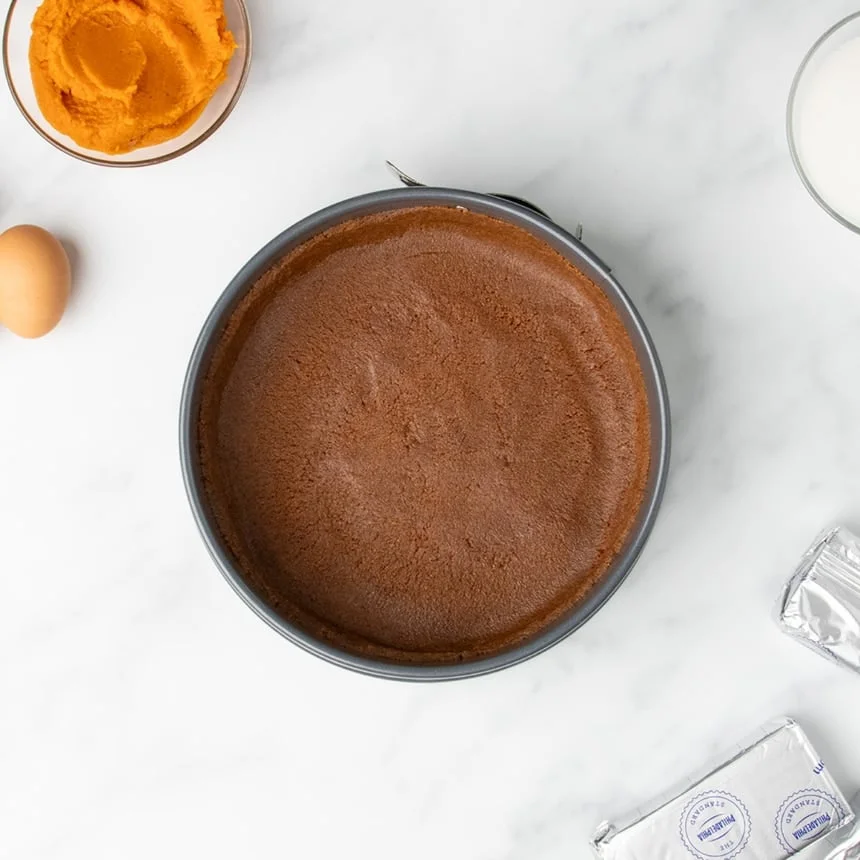 A springform pan with a dark, gluten free cookie crust pressed neatly inside, surrounded by other cheesecake ingredients on a marble counter.