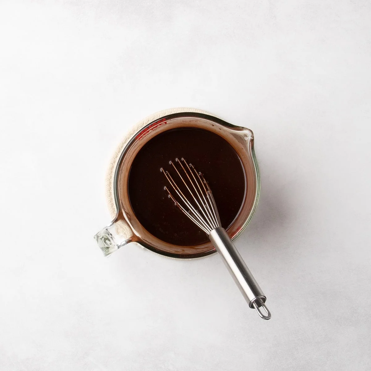 A whisk resting in a smooth dark chocolate mixture inside a glass measuring cup.