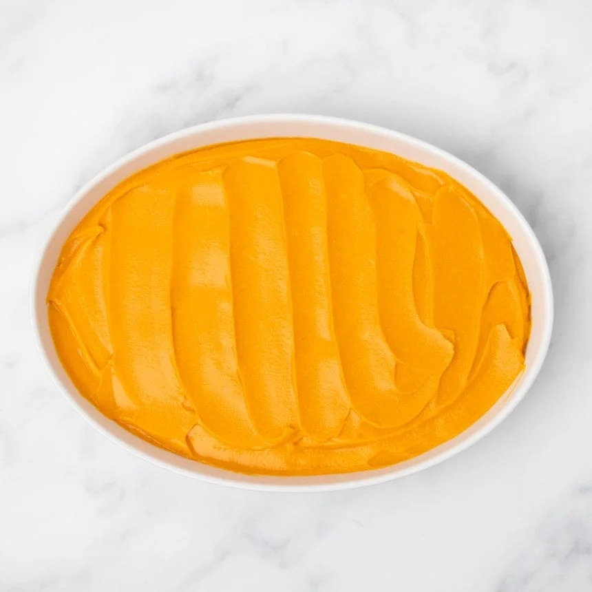 A white oval baking dish filled with evenly spread orange sweet potato filling on a white marble surface.