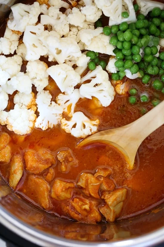 A wooden spoon stirring cauliflower and peas into tikka masala