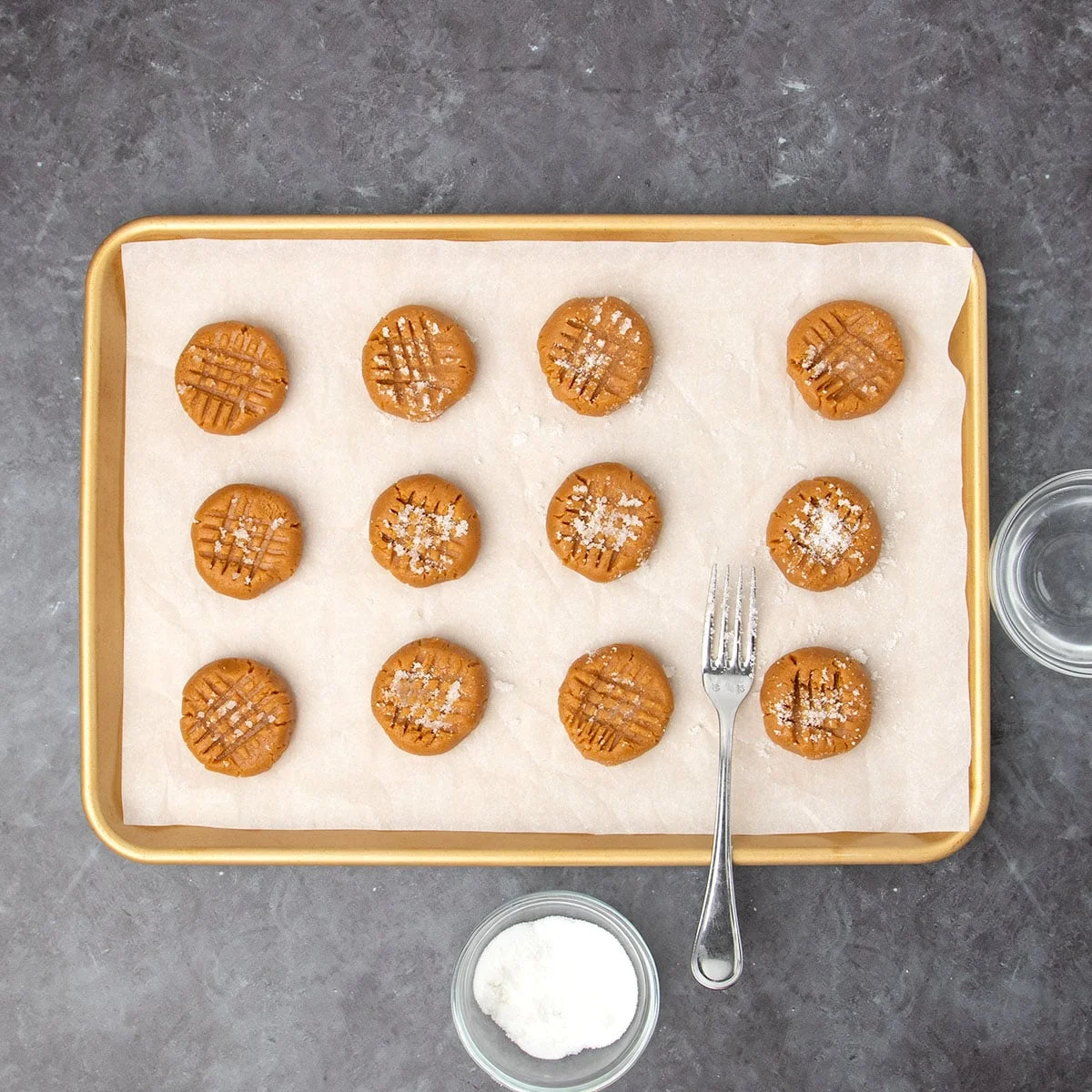 all 12 pieces of raw cookie dough with two crisscrossed marks from the fork tines with sugar on top on the same baking sheet