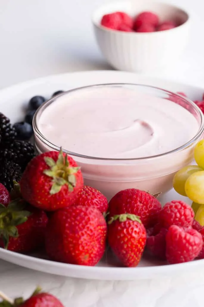 An assortment of fruit on a white plate with a glass bowl of pink fruit dip 