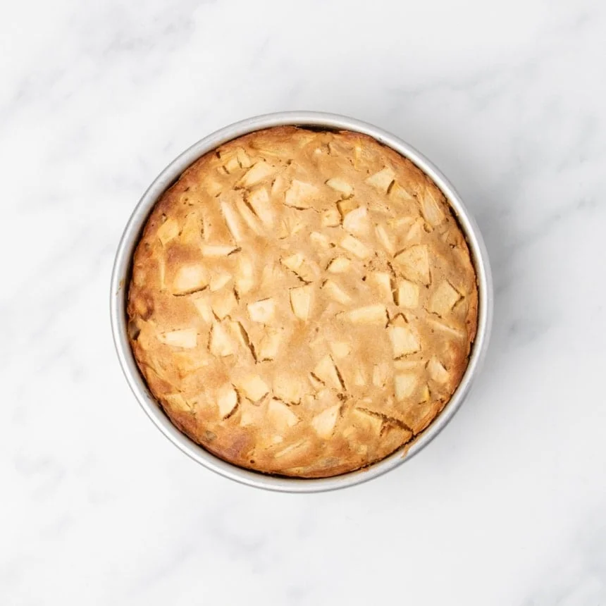 Baked apple cake in a round metal pan, golden brown on top with visible apple pieces.