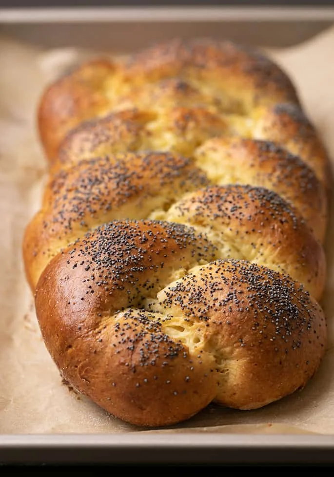 Baked braided challah bread with poppyseeds on top on brown paper lining metal baking pan