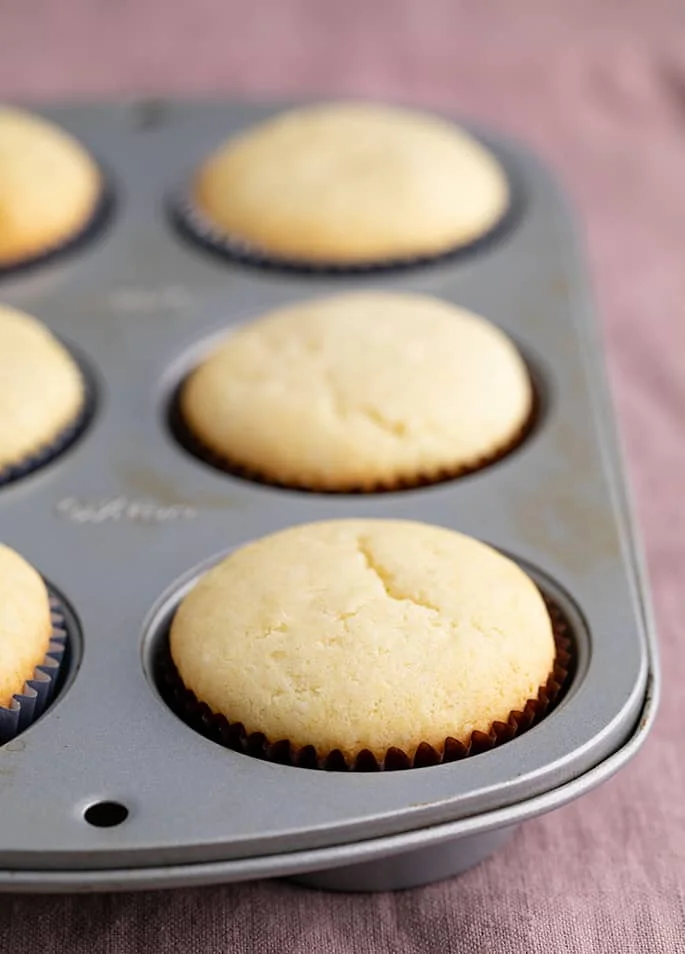 Baked gluten free vanilla cupcakes in 6 well cupcake tin in brown liners on purple cloth