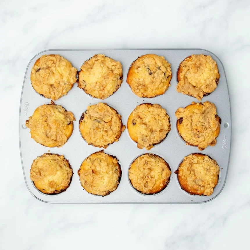 baked light brown muffins with golden brown crumble baked on top, still in metal muffin tin
