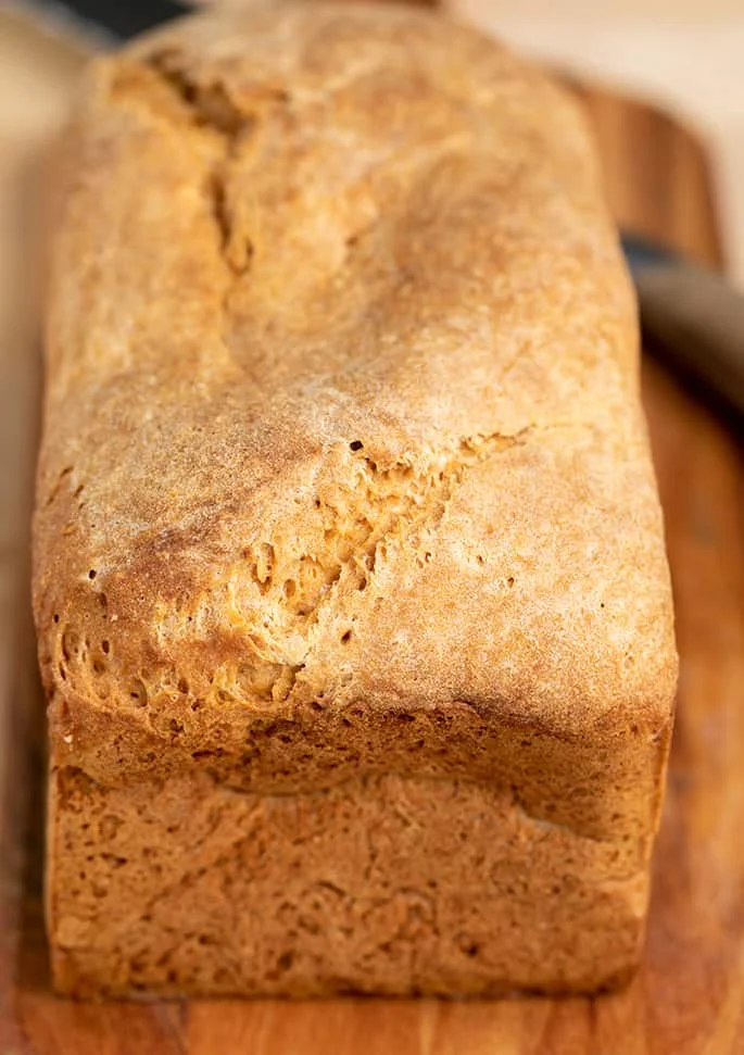 Baked whole loaf of potato bread on brown cutting board with bread knife.