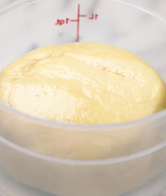 Ball of raw challah dough in 1 liter round plastic container on marble surface