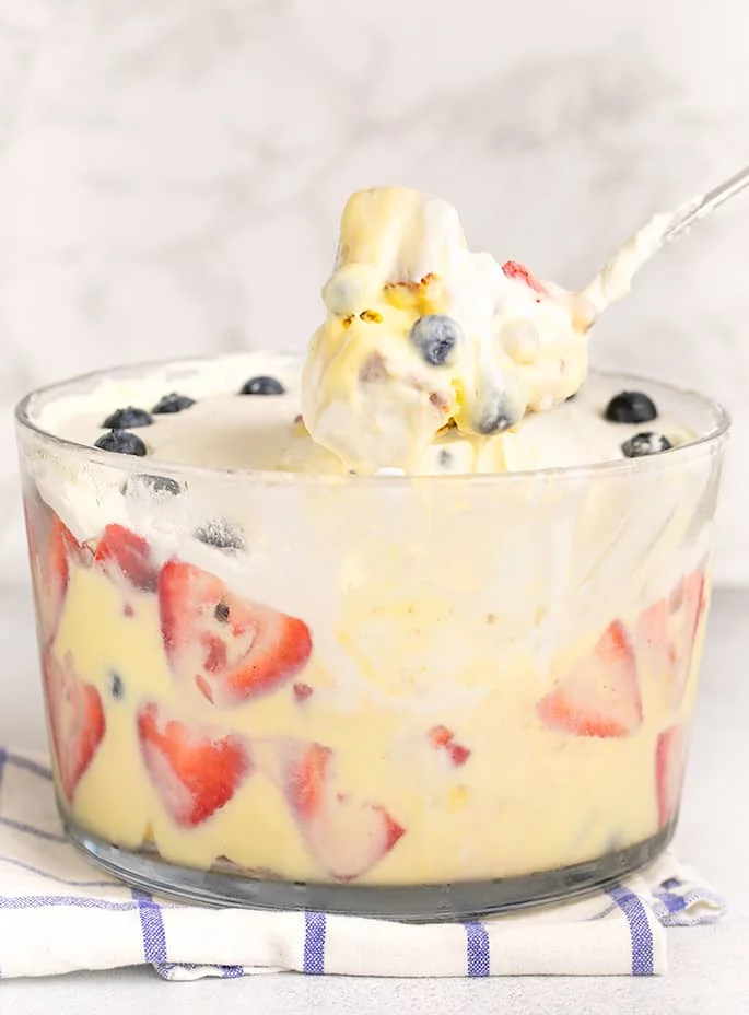 Berry custard trifle in glass bowl on blue and white windowpane cloth with spoonful on metal serving spoon