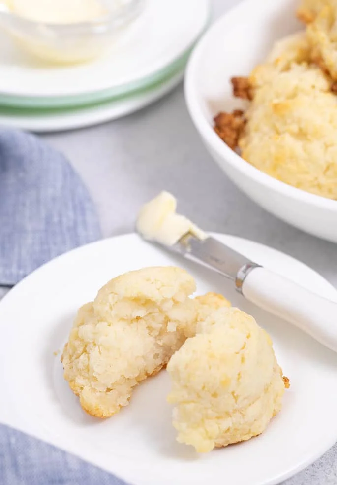 Biscuit split in half on small white plate with biscuits in bowl in background.
