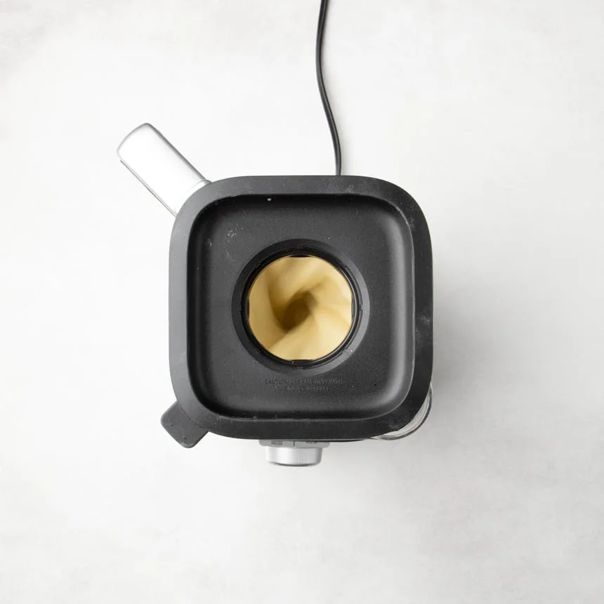 Blender with black lid on, showing light yellow batter blending again through the center opening.