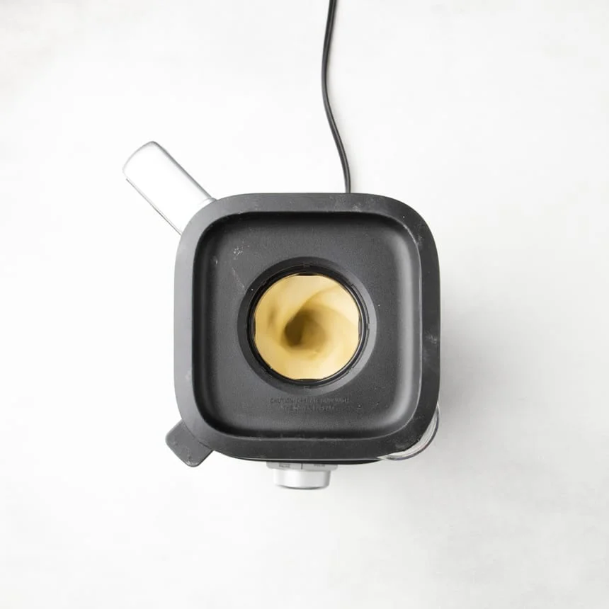 Blender with lid on, showing light yellow crepe batter blending through the center opening.