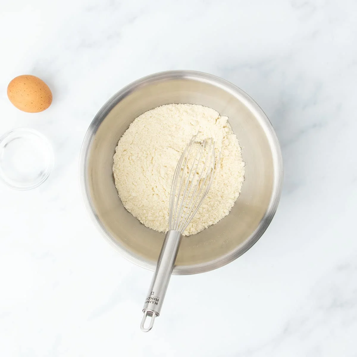 Bowl of almond flour with a whisk resting inside, egg and water nearby.