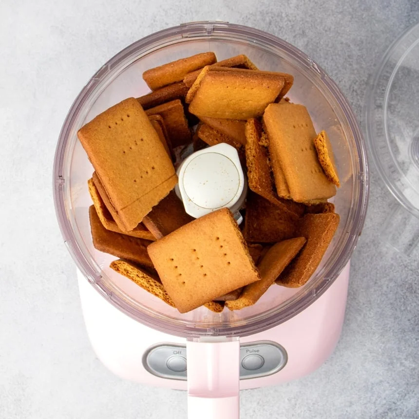 Broken brown gluten free graham crackers in pink colored food processor with open top.