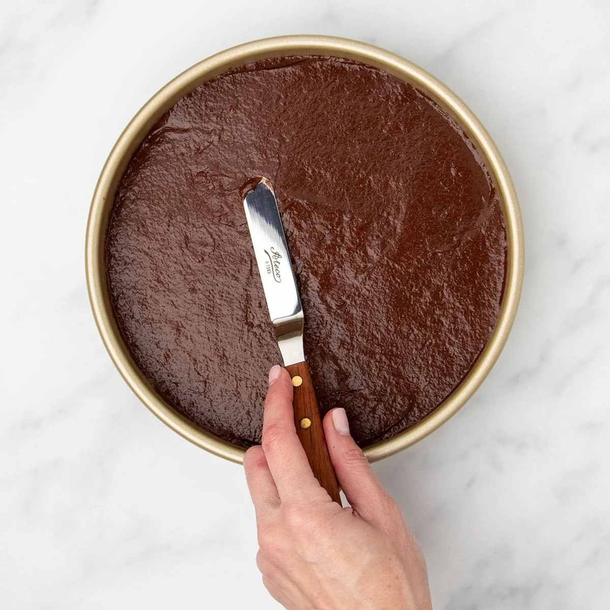 brown cake batter in round gold-colored baking pan with hand using small offset spatula to smooth top