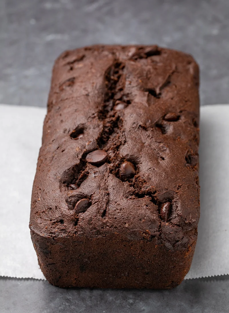 Brown loaf of cake with chocolate chips on slip of white paper on gray surface.