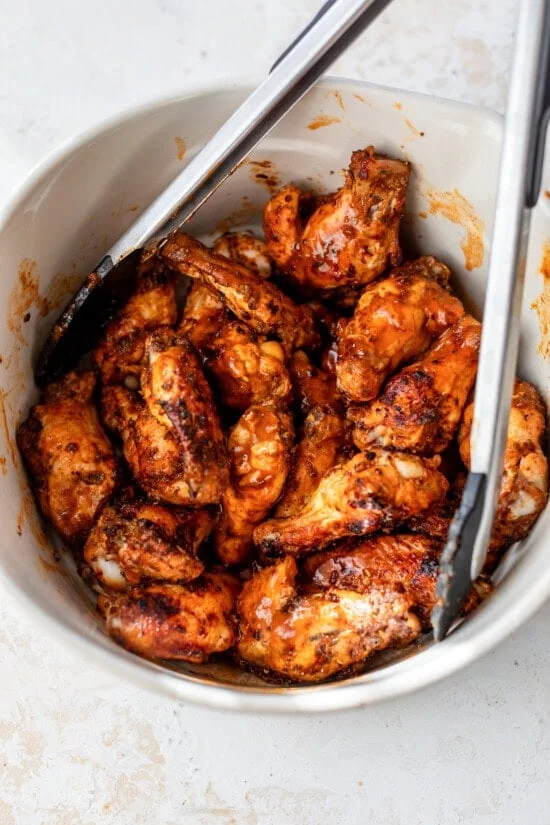 Buffalo chicken wings tossed in buffalo sauce in a bowl with tongs.