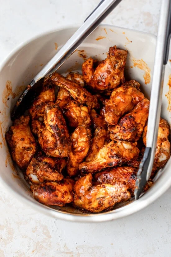 Buffalo chicken wings tossed in buffalo sauce in a bowl with tongs.