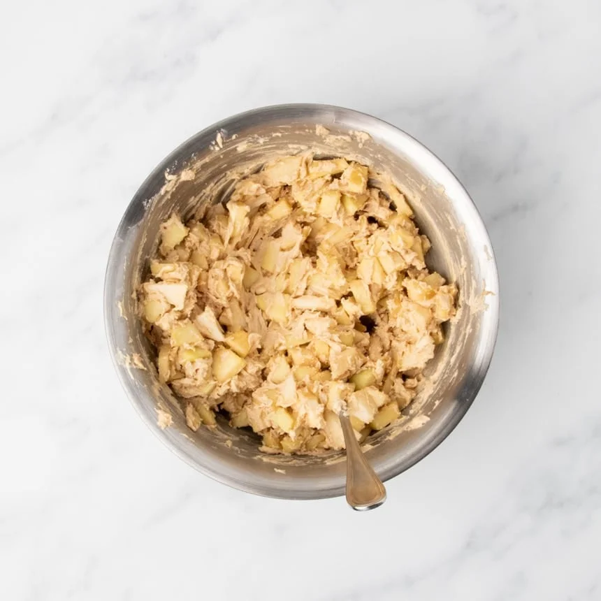 Cake batter mixed with diced apple pieces in a metal bowl with a spoon.