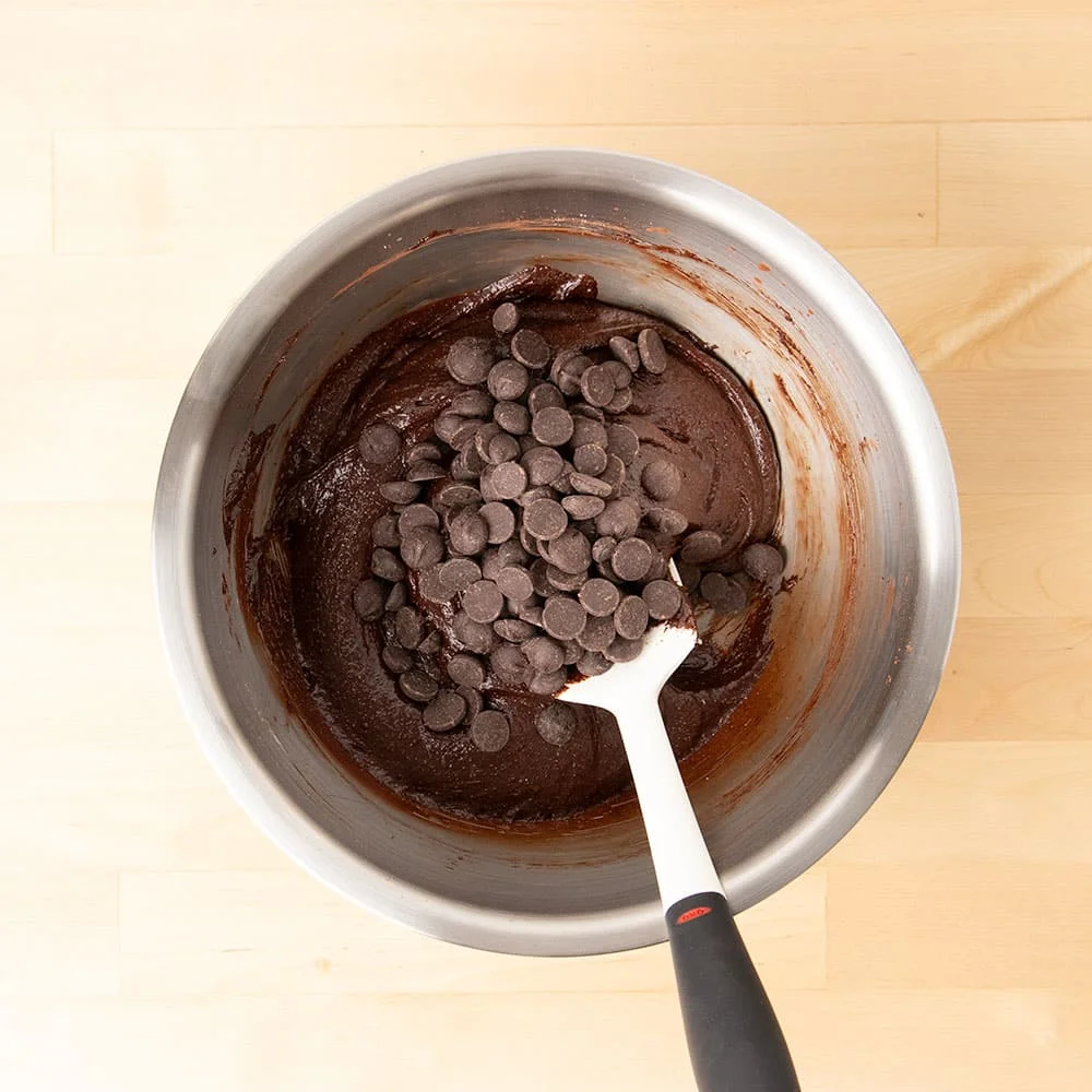 chocolate chips on top of dark brown brownie batter with white spatula in mixing bowl