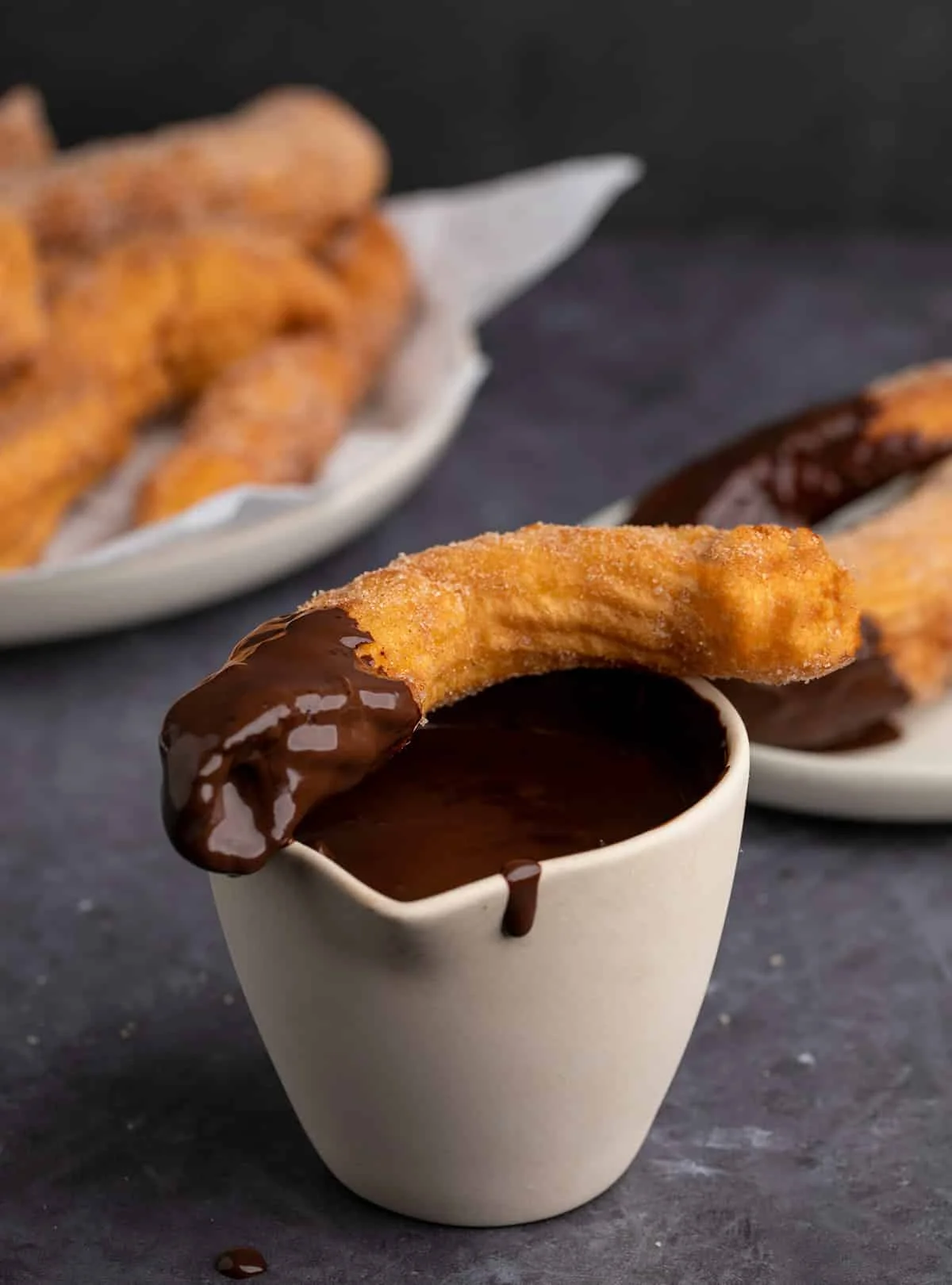 Churro dipped in chocolate sitting on jar of chocolate sauce with more churros in background