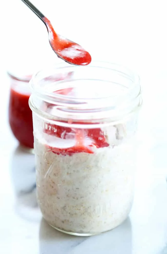 Close up of jar of overnight oats with spoon overhead with strawberry puree being poured in