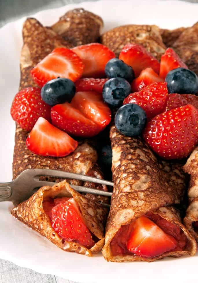 Close up of Swedish pancakes with fruit on top of them on a white plate being cut with a metal fork