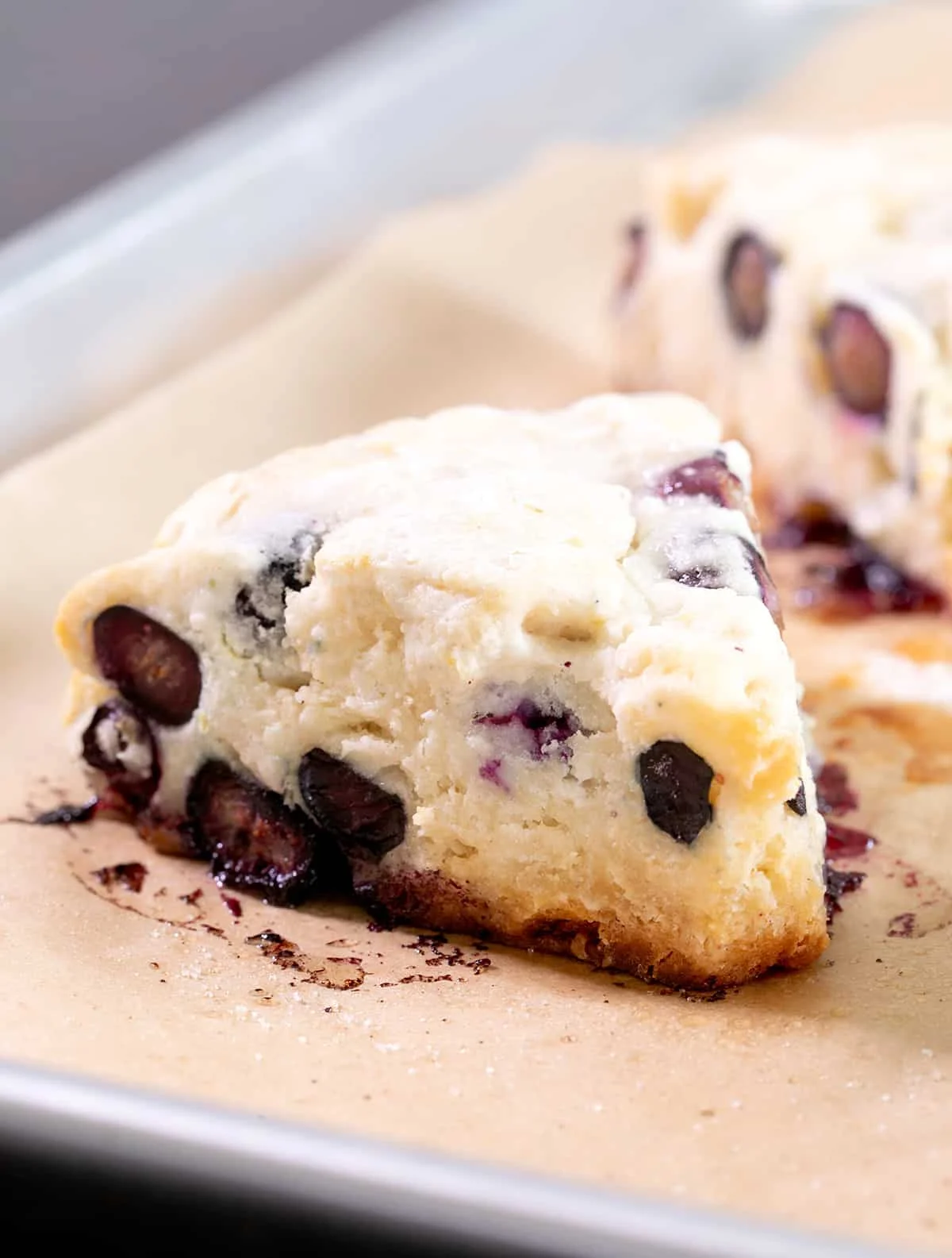 Closeup image of a light yellow baked triangle shaped pastry with blueberries on brown paper on a metal baking tray