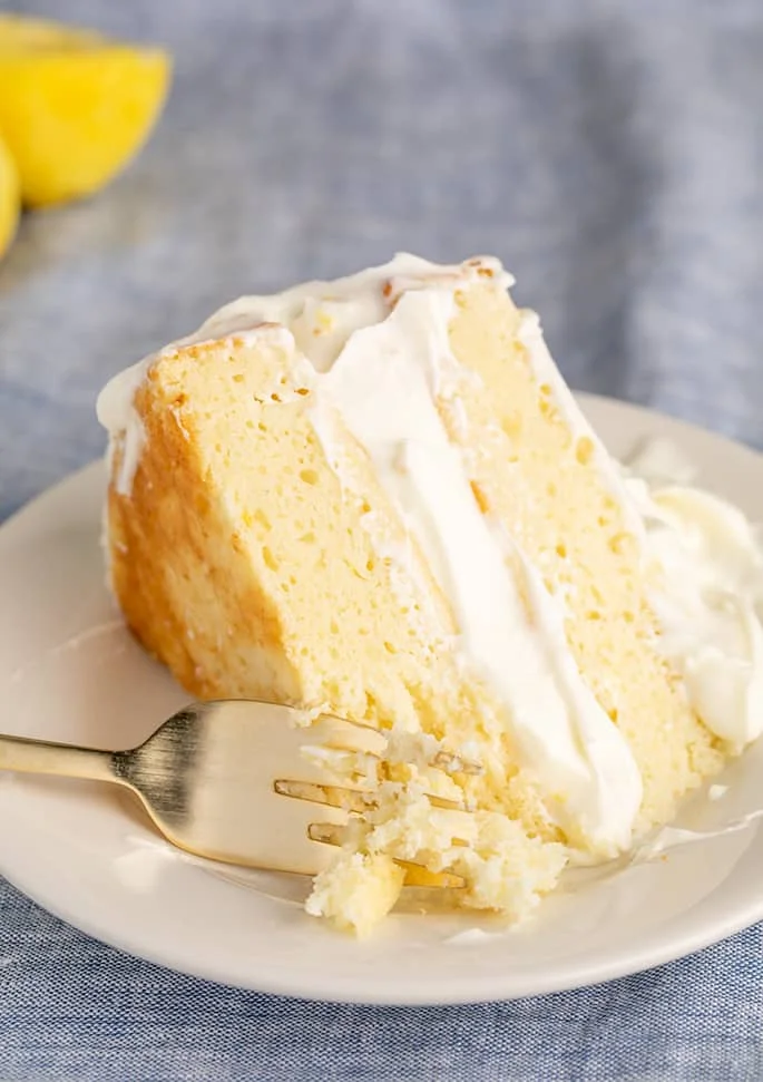 Closeup image of fork with cake crumbs on small white plate with slice of 2 layer lemon cake with lemon in background