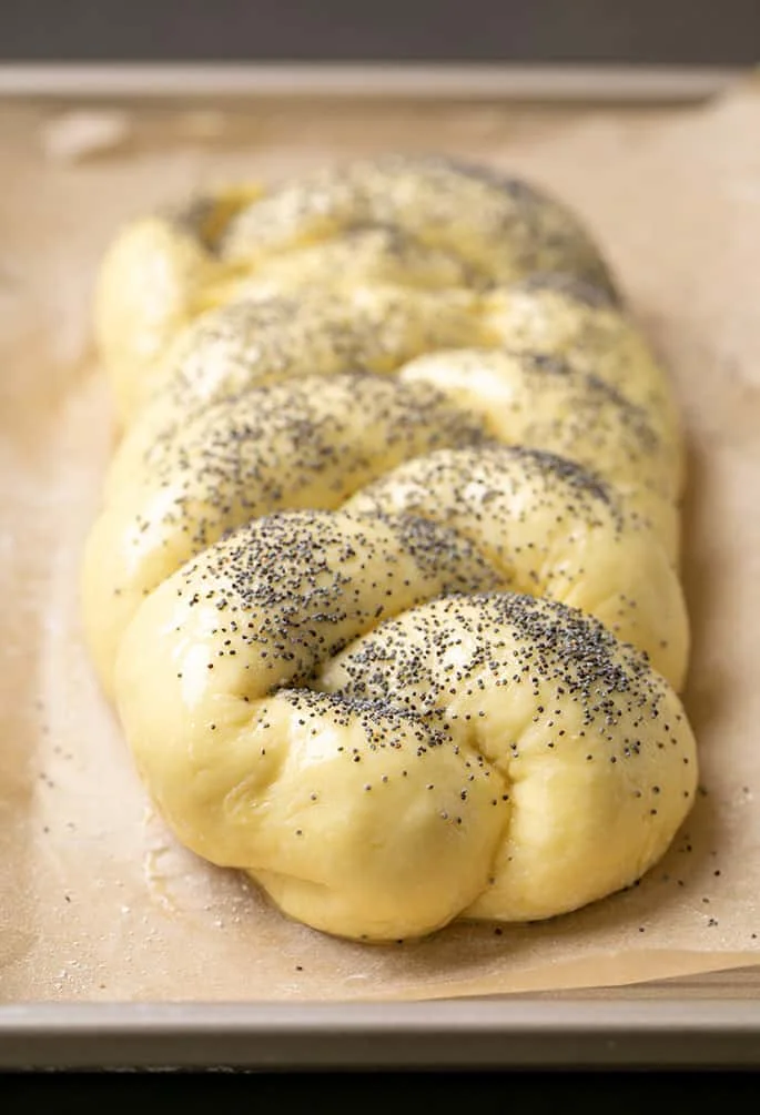Closeup image of raw braided challah dough with egg wash and poppyseeds on brown paper lining metal baking pan