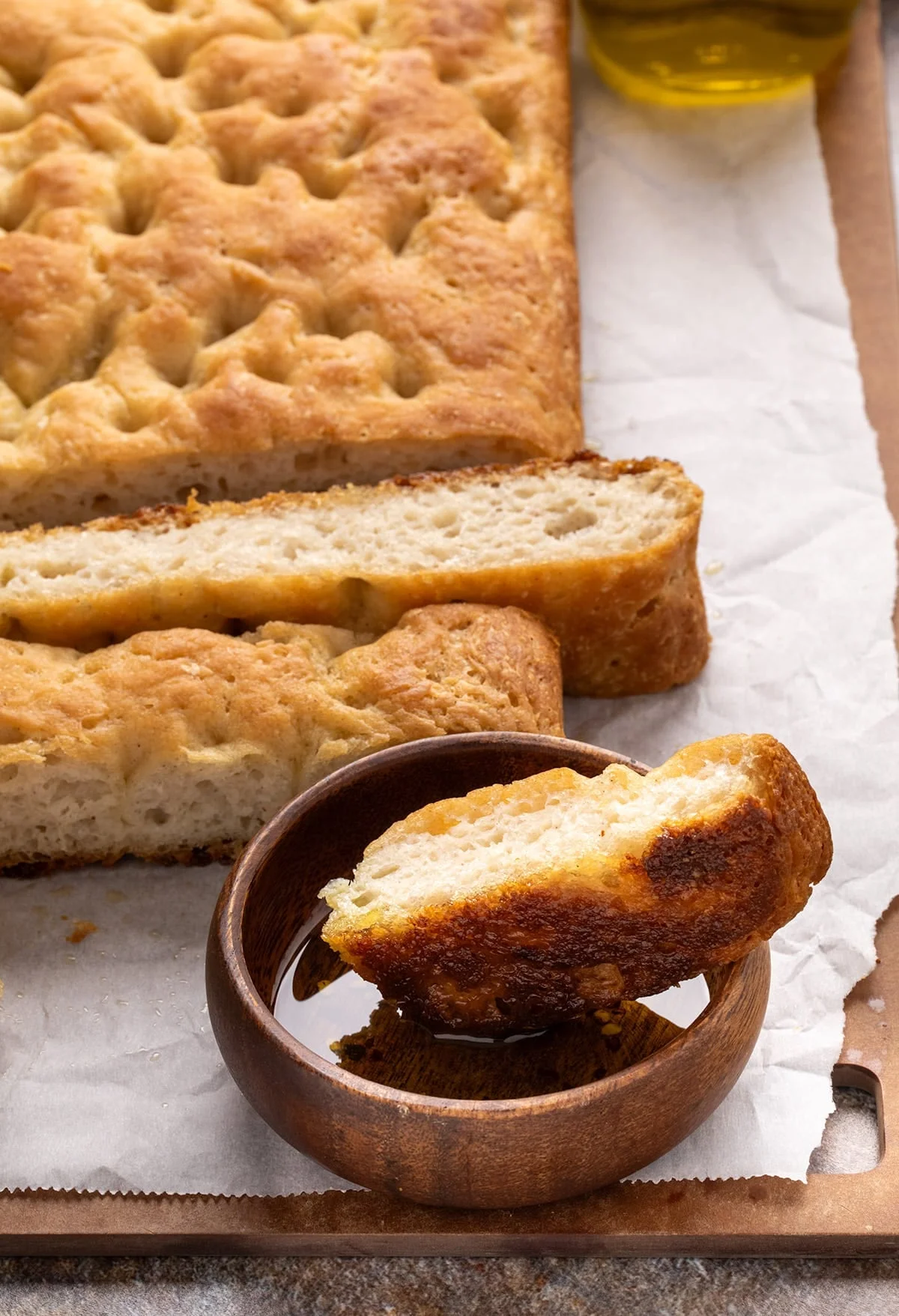 Closeup of a piece of bread dipped in oil inside a small wooden bowl, with more sliced focaccia visible in the background. The crust is deeply golden and crisp.