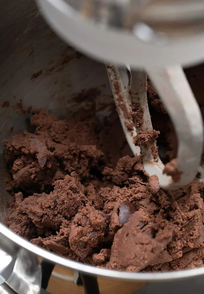 Clumps of brown cookie dough in a metal mixing bowl with paddle attachment